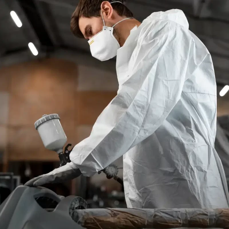 Un carrossier en tenue de protection et masque appliquant de la peinture sur la carrosserie d'une voiture.